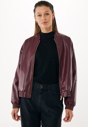 Giacca bomber in finta pelle bordeaux con zip dorata, polsini a costine e vestibilità comoda, indossata sopra un dolcevita nero e jeans neri.