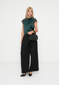 Dunkelgrünes Satinoberteil mit Spitzenakzenten, hoher Halsauschnitt, schwarze weite Hosen und eine strukturierte schwarze Handtasche. Schwarze spitze Absätze runden den Look ab.