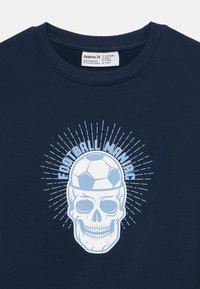 Σκούρο μπλε πουκάμισο με ένα κρανίο που φοράει μια μπάλα ποδοσφαίρου σαν καπέλο και το κείμενο «FOOTBALL MANIAC» τοξοειδώς από πάνω με ακτινωτές γραμμές.