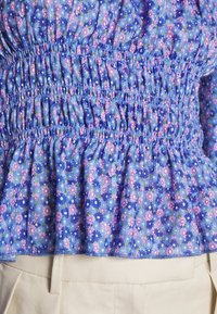 Bluse mit blauen Blumen und geraffter Textur, die pinke und weiße Blumen zeigt, mit elastischem Bund und Rüsche am Saum, kombiniert mit beigen Hosen.