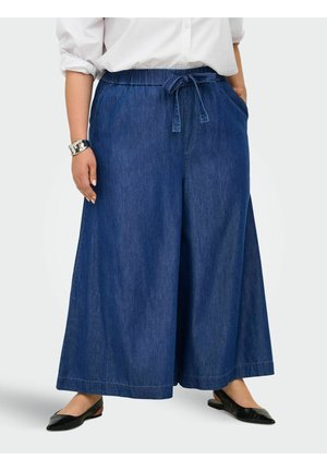 Femme portant un pantalon large en denim bleu avec une taille élastique et une ceinture nouée, associé à des ballerines noires et une chemise blanche.