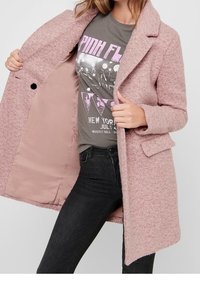 Rosa texturerad kappa med notsklapp och två framfickor, bärs över en grå t-shirt. Kappan har ett mjukt, ull-liknande material.