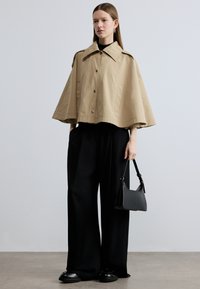 Bež vodootporna jakna-pelerina s velikim ovratnikom i srebrnim kopčama. Uparena s širokim crnim hlačama i crnom torbom preko ramena.