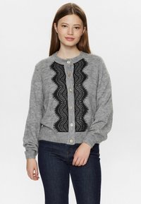 Šedý cardigan z měkkého materiálu s černými krajkovými detaily, kulatým výstřihem, zapínáním na knoflíky a dlouhými rukávy. Nositý přes tmavé džíny.