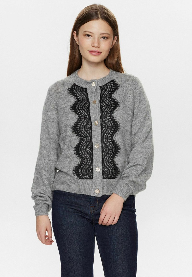 Šedý cardigan z měkkého materiálu s černými krajkovými detaily, kulatým výstřihem, zapínáním na knoflíky a dlouhými rukávy. Nositý přes tmavé džíny.