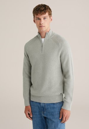 Hellgrauer, gerippter Strickpullover mit einem halblangen Reißverschlusskragen, langen Ärmeln und enganliegendem Saum, kombiniert mit blauen Jeans.