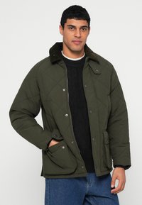 Casaco acolchoado verde-oliva com costura em forma de diamante, gola de veludo cotelê, dois bolsos com botão de pressão e fecho frontal de correr, usado sobre um suéter preto.