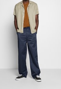 Långärmad beige skjorta med avslappnad passform, matchad med mörkblå vida byxor. Svarta sneakers fulländar looken och betonar den avslappnade stilen.