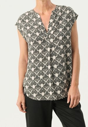 Femme portant un chemisier blanc sans manches avec des motifs symétriques de feuilles noires et un pantalon noir, debout devant un fond uni.