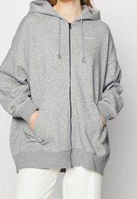 Haut à capuche zippé gris en tissu doux, doté d'une capuche, de poches kangourou et d'un logo Nike minimaliste sur la poitrine.