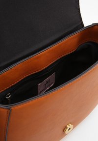 Brązowa torebka z ekoskóry z czarnym wnętrzem, wyposażona w zapięcie magnetyczne oraz małą kieszeń na zamek wewnątrz. Widoczna metka z logo.