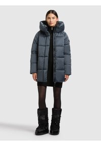 Žena u tamnoplatom prošivenom kaputu s visokim ovratnikom, crnoj pletenoj haljini, prozirnim čarapama i crnim zimskim čizmama, stoji na bijeloj pozadini.