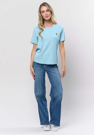 Kvinna som bär en ljusblå ribbad kortärmad topp med en liten broderad blomma, blå vida jeans och vita sneakers, stående mot en vit bakgrund.