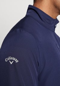 Marineblå genser med krage, laget av glatt materiale; har hvitt "Callaway"-logo og kontraststing på skulderen.