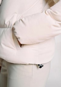 Mano nella tasca di un giubbotto a vento color crema con pantaloni abbinati, con un piccolo clip nero attaccato vicino alla vita.