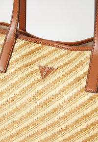 Sac fourre-tout en paille tressée avec poignées et finitions en cuir marron, orné d'une plaque triangulaire avec le logo Guess USA à l'avant.