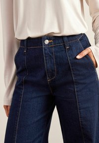Jean en denim bleu foncé avec une taille haute, présentant des coutures contrastées visibles, une fermeture à bouton sur le devant et des poches latérales.