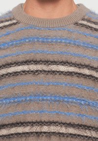 Close-up of a person wearing a beige knitted sweater with fuzzy horizontal stripes in blue, black, and white around the chest area.