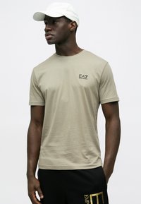 Világos khaki színű póló puha anyagból, rövid ujjakkal, kerek nyakkivágással és kis fekete "EA7" logóval. Fekete hozzáillő nadrággal viselve.