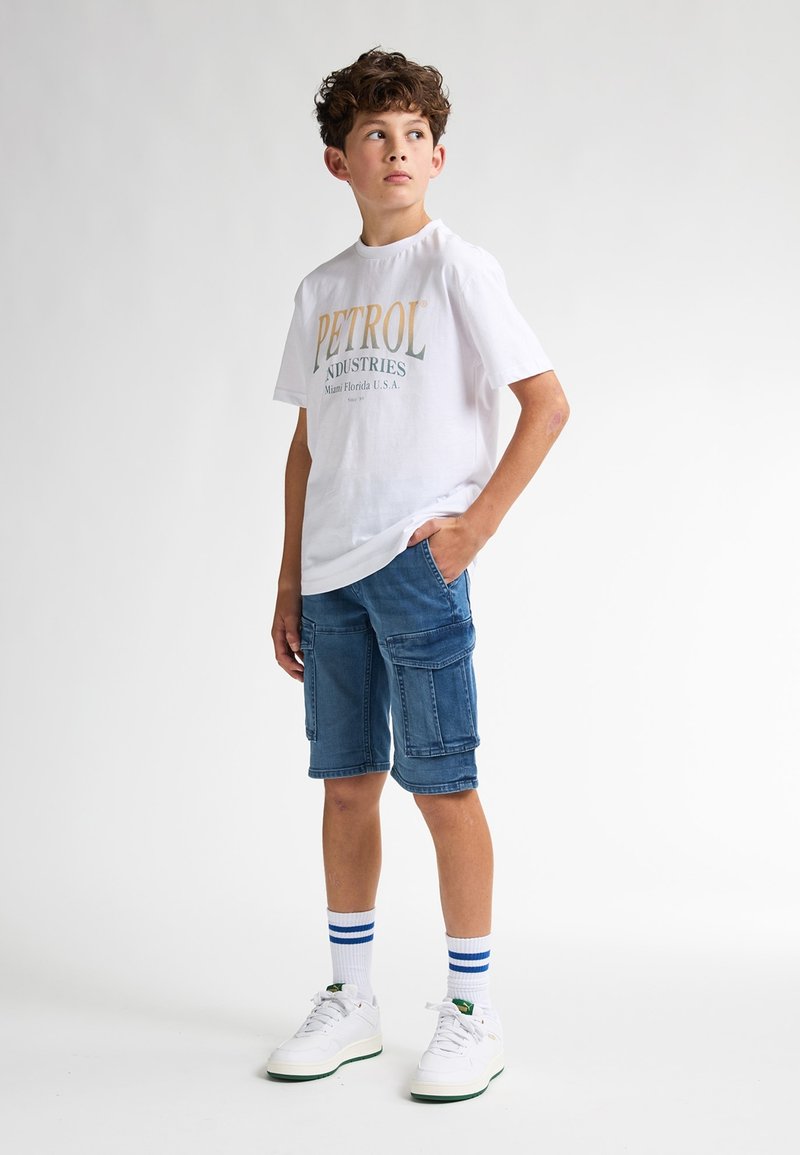 Camiseta blanca con el logo de "PETROL INDUSTRIES", pantalones cortos de cargo de mezclilla azul, zapatos blancos con acentos verdes y calcetines blancos con rayas azules.