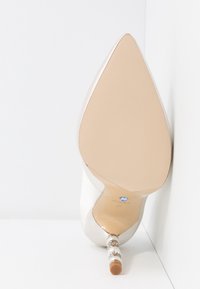 Kvinders spidse beige højhælede sko med dekorativ hæl med perler og rhinsten, set fra sålen mod en hvid hjørnebaggrund.