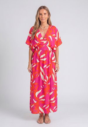 Femme portant une robe longue rose et orange à motifs, manches courtes, debout droite face à un fond clair uni.