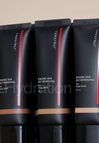 Tubes negros con un acabado brillante, franjas de acento rojas, etiquetados como "Synchro Skin Self-Refreshing Tint," que muestran SPF 20, en diferentes tonos de piel.