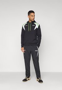Sort hætte sweatshirt med hvide og neongrønne detaljer, parret med sorte bukser med hvide striber og VENUM branding. Sko med et struktureret mønster.