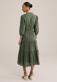 Abito verde oliva con maniche a sbuffo, design a strati e colletto arricciato. Il materiale sembra leggero e strutturato. Indossato con sandali wedges color sabbia.