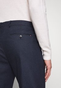 Pantaloni a quadri blu navy con un motivo sottile, dotati di una tasca posteriore con bottone e di una vita. Abbinati a un maglione in maglia di colore chiaro.