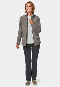 Lightweight, quilted grey jacket with a stand collar, zippered front, and two side pockets. Worn over a white top and paired with dark pants.