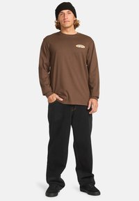 Camisa de manga larga marrón con parche de logo, combinada con pantalones negros holgados y zapatos negros. El portador lleva un gorro negro.
