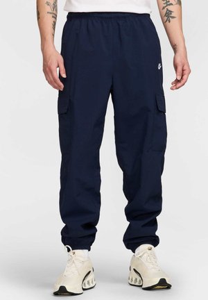 Pantalones cargo navy hechos de una tela ligera, con una cinturilla elástica, bolsillos laterales y puños. Combinados con zapatillas blancas.