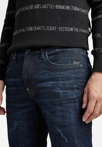 Los jeans de mezclilla azul oscura presentan un diseño clásico con un ligero desvanecimiento y arrugas. Una pequeña etiqueta de la marca es visible en el bolsillo trasero.