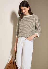 Pull en maille beige avec tricot aéré, poignets côtelés et un large décolleté, associé à un pantalon blanc à jambes larges et un sac fourre-tout en daim marron.