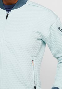 Světle modrá, strukturovaná bunda na zip s kulatým výstřihem, kontrastním tmavým límcem a zipovými kapsami. Na rameni je umístěno logo značky.