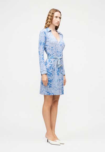 Γυναίκα που φοράει μακρυμάνικο μπλε φόρεμα με λευκά φύλλα και ζώνη στη μέση, στέκεται φορώντας λευκά μυτερά παπούτσια με χαμηλό τακούνι μπροστά σε απλό φόντο.