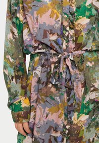 Rochie florală abstractă multicoloră cu mâneci lungi și curea din material legată în talie, având detalii cu volane de-a lungul placket-ului cu nasturi.