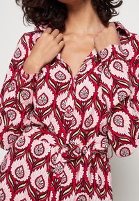 Czerwona sukienka koszulowa w kwiatowy wzór z długimi rękawami, zapinana na guziki, z pasującym paskiem; charakteryzuje się dużymi różowymi kwiatami z zielonymi akcentami.