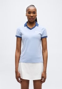 Γυναίκα που στέκεται και βλέπει μπροστά, φορώντας ανοιχτό μπλε πόλο μπλούζα με σκούρο μπλε γιακά και λευκή φούστα, μπροστά από ένα απλό φόντο.