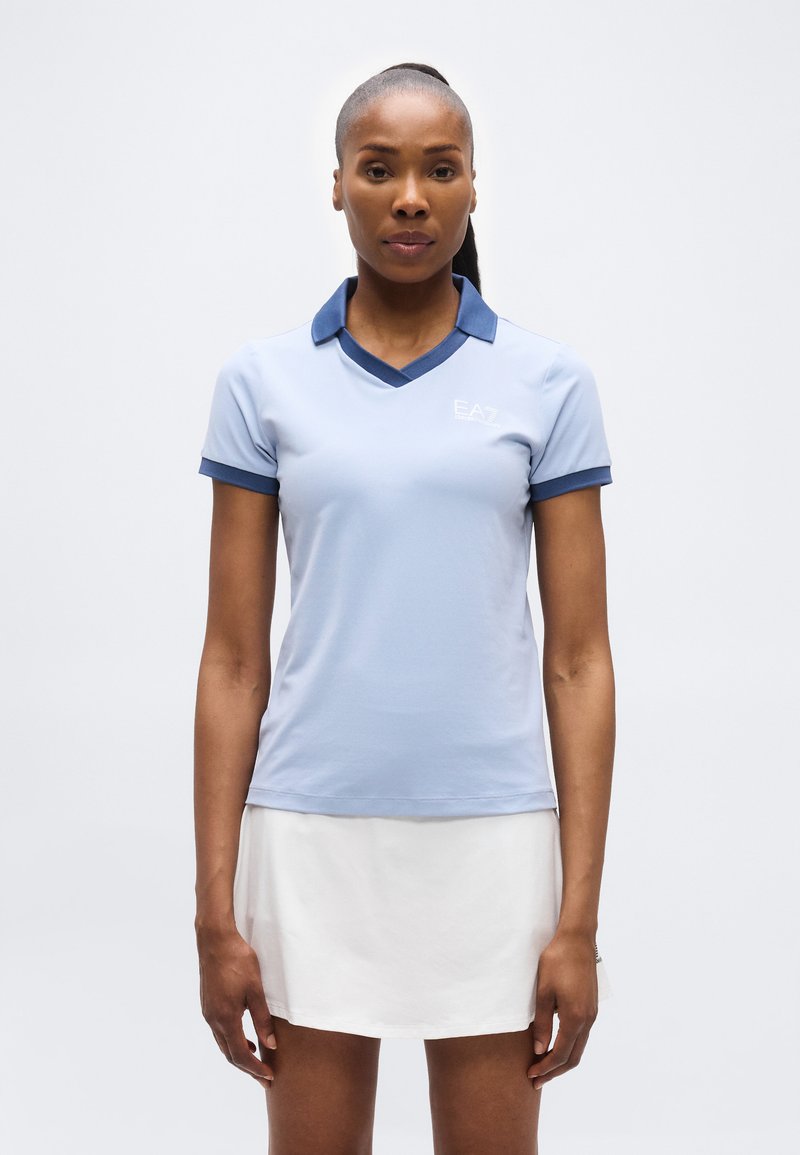 Γυναίκα που στέκεται και βλέπει μπροστά, φορώντας ανοιχτό μπλε πόλο μπλούζα με σκούρο μπλε γιακά και λευκή φούστα, μπροστά από ένα απλό φόντο.