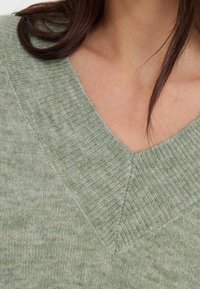 Pull vert clair en v fabriqué à partir d'un matériau en tricot texturé, avec un col côtelé et des motifs de couleur subtilement mélangés.