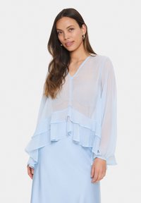 Blusa leggera azzurro chiaro, trasparente, con maniche lunghe, scollo a V e orlo a rouches sovrapposti. Tessuto liscio con una leggera lucentezza.