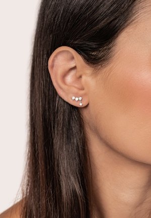 Nahaufnahme eines Ohrs mit drei kleinen silbernen und diamantenen Ohrringen, einer davon mit einem hängenden klaren Stein, vor glattem dunklem Haar und Haut.