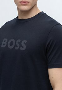 Gros plan d'une personne portant un T-shirt bleu marine foncé avec le mot BOSS imprimé en lettres légèrement plus foncées sur la poitrine.