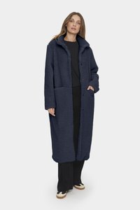 Mörkblå fleecejacka med en texturerad yta, oversized passform, stora fickor och krage, som matchas med svarta byxor och skor med beige accenter.
