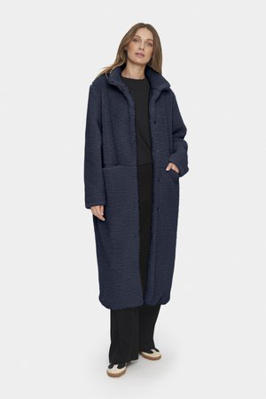 Cappotto in pile blu navy con finitura testurizzata, vestibilità oversize, grandi tasche e colletto, abbinato a pantaloni neri e scarpe con dettagli beige.