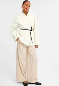 Vit, överdimensionerad kimono-inspirerad jacka med ett svart bälte, kombinerad med vida beiga byxor och svarta loafers. Slät tyg, minimalistisk design.