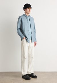 Cămașă cu dungi albastre deschise, cu butoane, purtată împreună cu un tricou albastru uniform, pantaloni albi și pantofi negri; modelul stă în fața unui fundal neutru.