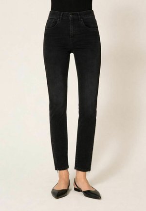 Jeans neri slim fit a vita alta con dettagli a zip alla caviglia, indossati con ballerine slingback nere con punta a punta, su uno sfondo neutro.